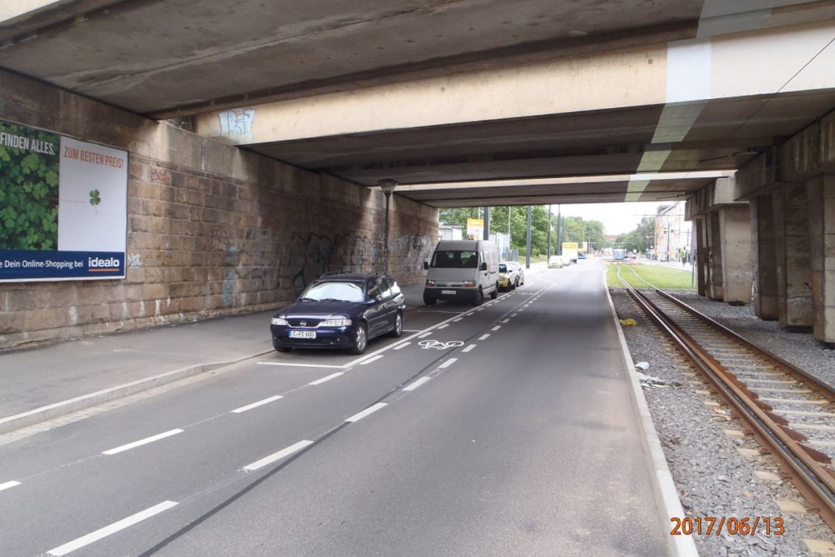 Unter einer Brücke sind von Links nach rechts ein Fußweg, parkende Autos, ein Radweg und dann Schienen für einen Zug oder eine Straßenbahn