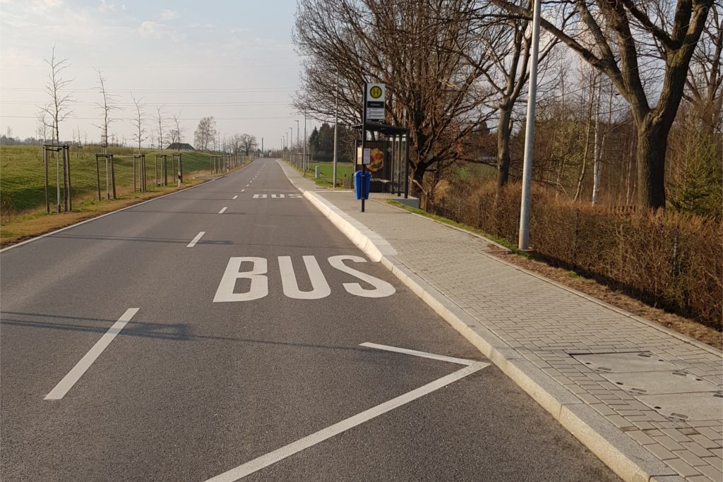 Eine Straße läuft vom Vordergrund gerade in den Hintergrund, auf der rechten Seite hat sie eine Haltestelle für Buss