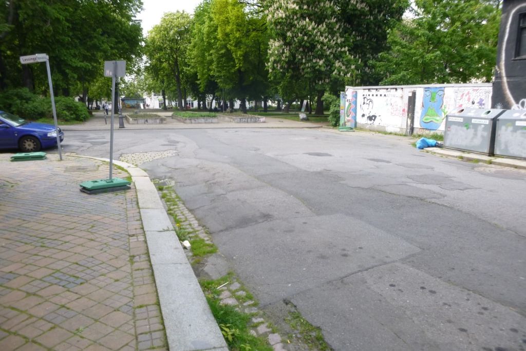 Im Hintergrund sieht man den Lessingplatz in Chemnitz, welcher von Bäumen umgeben ist und Betonierte große Blumenkästen in der Mitte hat. Zum Lessingplatz hin, führt eine Straße